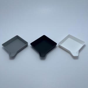 Può includere: Tre piccoli vassoi quadrati in fila, ciascuno con un beccuccio su un lato. I vassoi sono in tonalità di grigio, nero e bianco. I vassoi vengono probabilmente utilizzati per contenere piccoli oggetti o per servire.