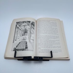 May include: An open book with illustrations and text, held open by a black book stand. The book's pages are yellowed, and the text is in black. The illustration depicts a wooden structure with a long table.