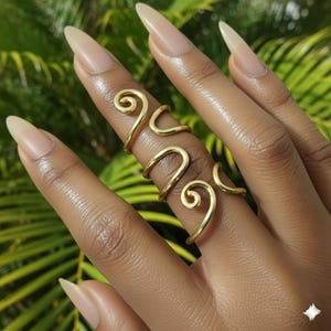 May include: Gold-colored, adjustable ring with a unique, swirling design. The ring wraps around the finger with a series of curved lines and spiral accents. The jewelry is displayed on a hand with long, polished nails.