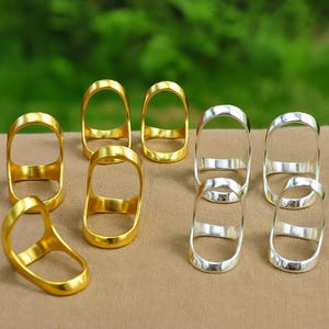 以下が含まれることがあります： 金と銀の指輪のコレクション。リングは、指を包むバンドが付いた楕円形のデザインです。金のリングは光沢のある仕上がりで、銀のリングは磨かれた外観です。リングはベージュ色の表面に表示されています。