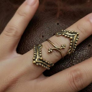 May include: An antique gold-colored finger ring with a unique design. The ring features two ornate, square-shaped elements connected by a delicate, curved band. The square elements are adorned with small, raised dots, adding texture and detail to the jewelry.