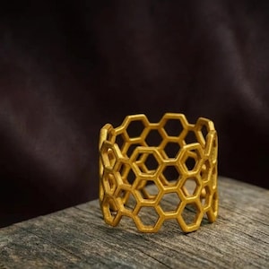 May include: A gold-coloured napkin ring featuring a repeating hexagonal honeycomb pattern. The structure is composed of interconnected geometric shapes forming a cylindrical band with a textured, matte finish throughout the entire object.