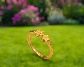 May include: A gold-coloured ring with two star-shaped features. The ring is set against a blurred background of green grass and colourful flowers. The ring's design is simple and elegant.