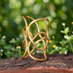 May include: A gold-toned cuff bracelet featuring an openwork design of interconnected, fluid, organic loops. The structure is composed of smooth, rounded metal lines that create a cage-like appearance. The text Prao Jewels is visible.