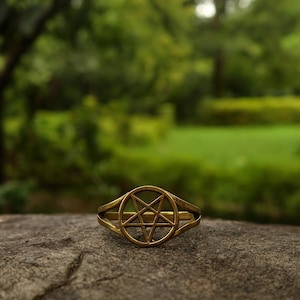 May include: A gold-colored ring featuring a pentagram design within a circular frame. The ring is set against a blurred green background, suggesting an outdoor setting. The ring's design is the focal point.