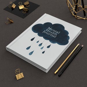 May include: A white hardcover journal featuring a dark blue watercolor cloud graphic with the text Mental Health Journal printed in white, accompanied by several blue watercolor raindrops falling from the cloud onto the cover surface.