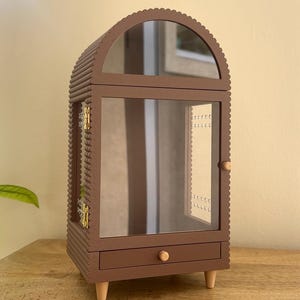 Pode incluir: Um porta-joias castanho com um exterior texturizado e canelado com um topo arqueado. Possui uma porta frontal de vidro com um pequeno puxador de madeira, uma sec&ccedil;&atilde;o superior espelhada, uma gaveta inferior com puxador e quatro pernas curtas.