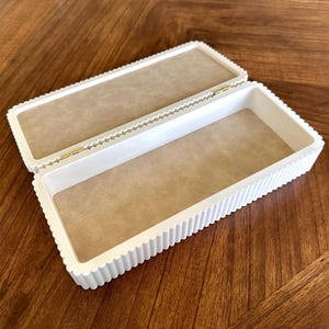 K&ouml;nnte beinhalten: Eine offene, rechteckige wei&szlig;e Box mit geriffelter Au&szlig;enseite und beigefarbenem Innenfutter. Die Box ist mit goldfarbener Hardware versehen. Die Box steht auf einer Holzoberfl&auml;che.