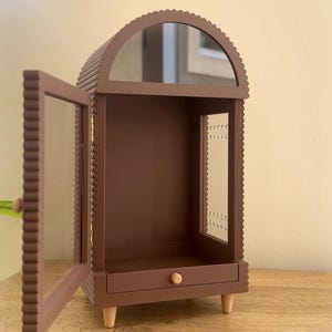 Pode incluir: Uma vitrine arqueada castanha com uma moldura recortada e uma porta de vidro aberta. O m&oacute;vel possui uma pequena gaveta com um puxador de madeira redondo na base, uma sec&ccedil;&atilde;o superior espelhada e quatro pernas de madeira clara.