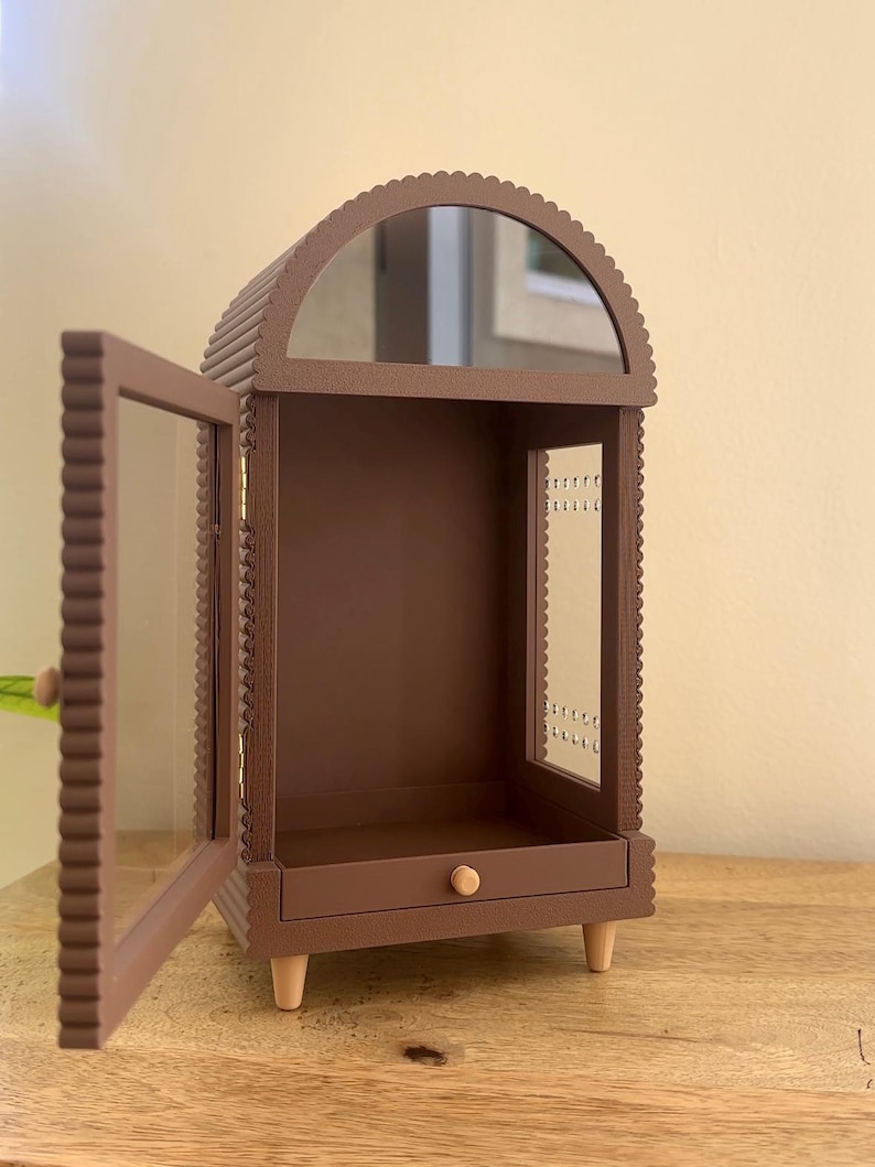 Pode incluir: Uma vitrine arqueada castanha com uma moldura recortada e uma porta de vidro aberta. O m&oacute;vel possui uma pequena gaveta com um puxador de madeira redondo na base, uma sec&ccedil;&atilde;o superior espelhada e quatro pernas de madeira clara.
