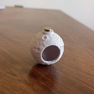 Puede incluir: Objeto esférico blanco con forma de luna, con superficie texturizada y abertura circular. Tiene una pequeña tapa metálica. Probablemente sea un objeto decorativo o un recipiente.