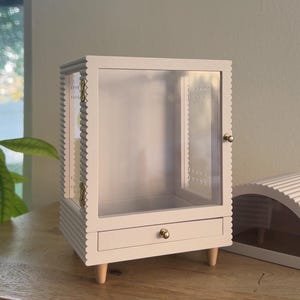 Pode incluir: Uma vitrine branca com moldura recortada, porta frontal transparente e uma gaveta &uacute;nica com um pequeno puxador dourado. O m&oacute;vel &eacute; sustentado por quatro p&eacute;s de madeira clara e inclui dobradi&ccedil;as de metal dourado na porta lateral.
