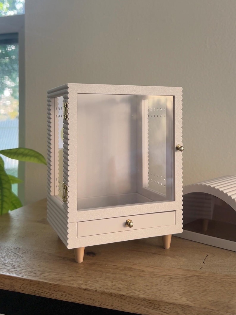 Pode incluir: Uma vitrine branca com moldura recortada, porta frontal transparente e uma gaveta &uacute;nica com um pequeno puxador dourado. O m&oacute;vel &eacute; sustentado por quatro p&eacute;s de madeira clara e inclui dobradi&ccedil;as de metal dourado na porta lateral.