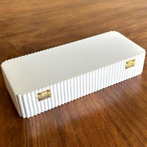 K&ouml;nnte beinhalten: Eine rechteckige, cremefarbene Box mit geriffeltem Design und goldfarbenen Metallscharnieren. Die Box ist geschlossen und liegt auf einer Holzoberfl&auml;che.
