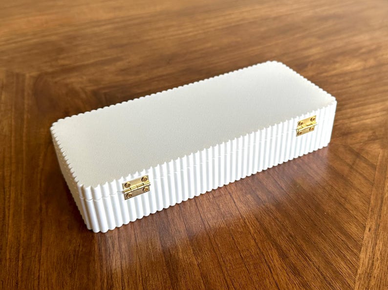 K&ouml;nnte beinhalten: Eine rechteckige, cremefarbene Box mit geriffeltem Design und goldfarbenen Metallscharnieren. Die Box ist geschlossen und liegt auf einer Holzoberfl&auml;che.