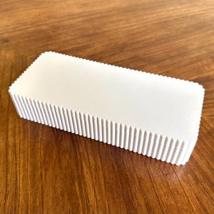 K&ouml;nnte beinhalten: Rechteckige, cremefarbene Box mit geriffeltem, strukturiertem Design an den Kanten. Die Box steht auf einer warmen, braunen Holzoberfl&auml;che, was einen Kontrast erzeugt. Das Objekt scheint ein Beh&auml;lter oder eine Verpackung zu sein. Ca. 15 cm x 7 cm.
