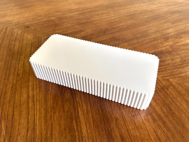K&ouml;nnte beinhalten: Rechteckige, cremefarbene Box mit geriffeltem, strukturiertem Design an den Kanten. Die Box steht auf einer warmen, braunen Holzoberfl&auml;che, was einen Kontrast erzeugt. Das Objekt scheint ein Beh&auml;lter oder eine Verpackung zu sein. Ca. 15 cm x 7 cm.