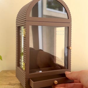 Pode incluir: Um porta-joias castanho com moldura recortada, topo arqueado e porta de vidro transparente. O m&oacute;vel possui uma pequena gaveta na base com um puxador redondo, dobradi&ccedil;as de metal dourado e pain&eacute;is laterais internos com perfura&ccedil;&otilde;es.