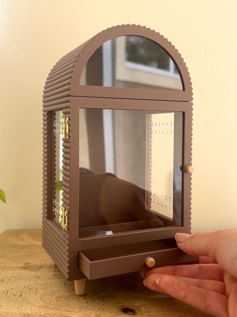 Pode incluir: Um porta-joias castanho com moldura recortada, topo arqueado e porta de vidro transparente. O m&oacute;vel possui uma pequena gaveta na base com um puxador redondo, dobradi&ccedil;as de metal dourado e pain&eacute;is laterais internos com perfura&ccedil;&otilde;es.
