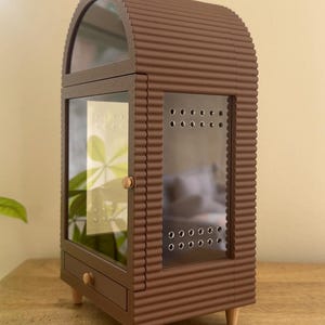 Pode incluir: Uma vitrine castanha em forma de arco com textura exterior canelada, apresentando uma porta de vidro com orif&iacute;cios de ventila&ccedil;&atilde;o, uma pequena gaveta com um puxador redondo e quatro pernas de madeira clara e c&oacute;nicas. A vitrine tem um painel frontal.