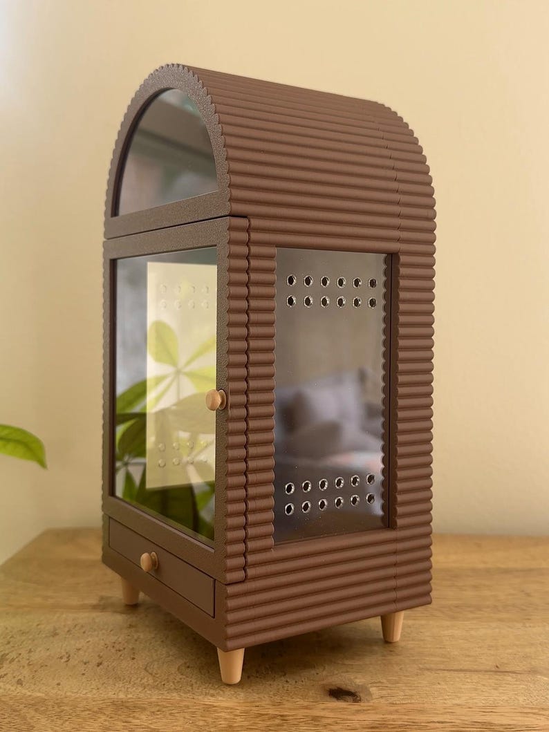 Pode incluir: Uma vitrine castanha em forma de arco com textura exterior canelada, apresentando uma porta de vidro com orif&iacute;cios de ventila&ccedil;&atilde;o, uma pequena gaveta com um puxador redondo e quatro pernas de madeira clara e c&oacute;nicas. A vitrine tem um painel frontal.