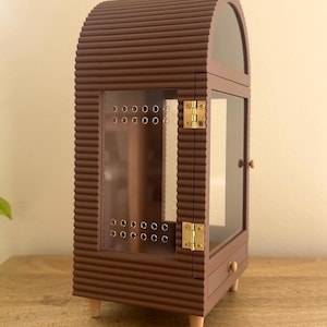 Pode incluir: Uma vitrine arqueada castanha com uma textura exterior canelada. Possui uma porta frontal transparente com dobradi&ccedil;as de metal dourado, um pequeno puxador redondo e uma s&eacute;rie de orif&iacute;cios de ventila&ccedil;&atilde;o. O m&oacute;vel assenta em quatro pernas curtas claras.