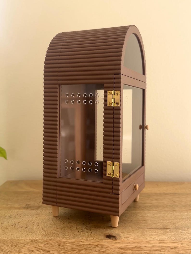 Pode incluir: Uma vitrine arqueada castanha com uma textura exterior canelada. Possui uma porta frontal transparente com dobradi&ccedil;as de metal dourado, um pequeno puxador redondo e uma s&eacute;rie de orif&iacute;cios de ventila&ccedil;&atilde;o. O m&oacute;vel assenta em quatro pernas curtas claras.