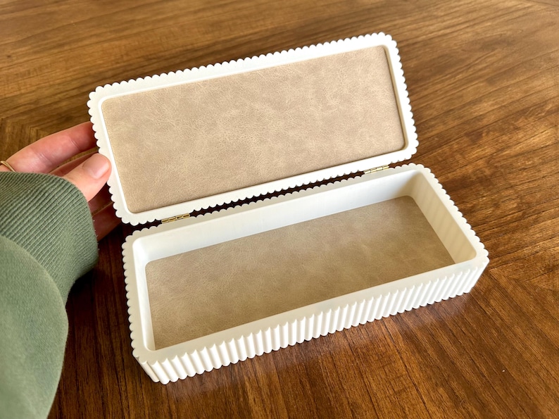 K&ouml;nnte beinhalten: Ge&ouml;ffnete wei&szlig;e rechteckige Box mit einem gewellten Randdesign. Der Innenraum ist mit einem beigefarbenen Stoff ausgekleidet. Die Box steht auf einer Holzoberfl&auml;che.