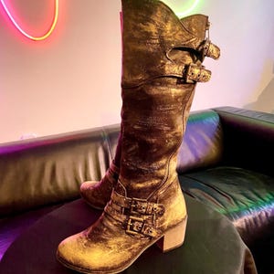 Peut inclure: Bottes hautes au genou avec une finition dorée vieillie. Elles comportent plusieurs fermetures à boucles et un talon épais. La matière des bottes a un aspect texturé. Les bottes sont présentées sur un fond neutre.