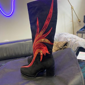 May include: Black suede knee-high boots with a decorative red and gold sequined flame design. The boots have a block heel and platform sole. A stylish and unique footwear choice.