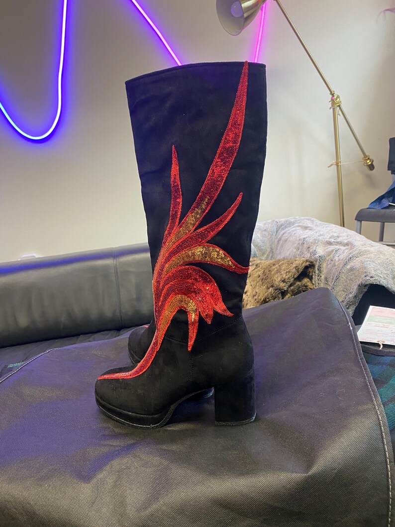 May include: Black suede knee-high boots with a decorative red and gold sequined flame design. The boots have a block heel and platform sole. A stylish and unique footwear choice.
