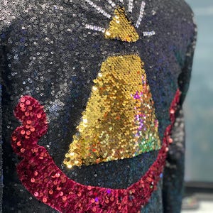 Peut inclure: Veste noire à paillettes avec un motif doré et rouge à paillettes dans le dos. Le motif présente un triangle doré avec un éclat blanc au-dessus et un accent rouge incurvé à paillettes en dessous.