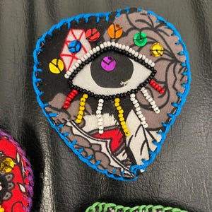 May include: Heart-shaped fabric patch with a detailed eye design. The patch features a black background with a gray and white pattern, a white eye, and colorful sequin accents. The edges are outlined with blue stitching.