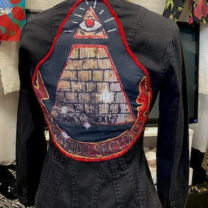 Peut inclure: Veste en jean noire avec un design unique au dos. La veste présente un écusson avec un œil dans un triangle, une pyramide et les mots "ORDO SECLORUM" en lettres dorées. Le col a un motif léopard.