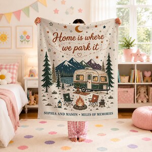 May include: A cream-colored throw blanket featuring a printed camping scene with a vintage camper, mountains, pine trees, a campfire, and chairs. The text reads "Home is where we park it" and "SOPHIA AND MASON • MILES OF MEMORIES" in brown.