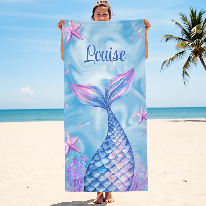 May include: A blue beach towel with a mermaid tail design. The towel features a watercolor illustration of a mermaid tail in shades of blue and pink, with the name "Louise" in script. Pink starfish and bubbles are also included.
