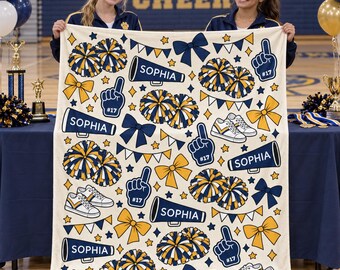 May include: A cream-colored fleece blanket featuring a repeating pattern of navy blue and gold cheerleading motifs, including pom-poms, bows, sneakers, foam fingers labeled #17, and megaphones with the name Sophia printed in white text.