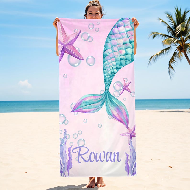 May include: A pink beach towel featuring a mermaid tail design in shades of blue and purple, with starfish and bubbles. The name "Rowan" is written in cursive at the bottom. The towel is held up on a beach.