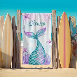 May include: A beach towel with a mermaid tail design in shades of teal, purple, and blue, with the name "Elowen" in script. The towel is displayed on a wooden fence with surfboards on either side, set against a beach and ocean backdrop.