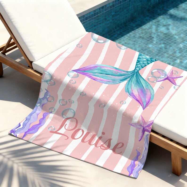 May include: A pink and white striped beach towel featuring a mermaid tail in shades of blue and purple, bubbles, and starfish. The name "Louise" is written in cursive. The towel is draped over a white lounge chair.