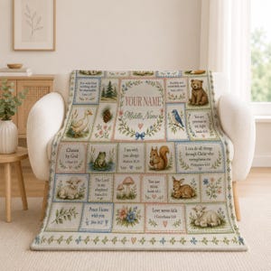 May include: A cream-colored throw blanket featuring a grid of rectangular panels with illustrations of a bear, duck, frog, squirrel, fawn, and lambs, interspersed with floral wreaths and Bible verses, including the text YOUR NAME Middle Name.