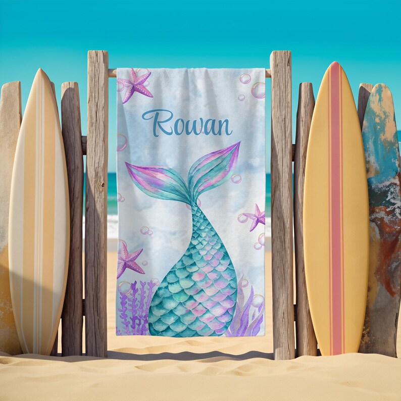 May include: A personalized beach towel featuring a mermaid tail design in shades of teal, purple, and pink. The name "Rowan" is written in blue script. The towel is displayed on a wooden fence with surfboards on either side.