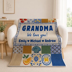 May include: A soft throw blanket featuring a cream-colored center with the blue text GRANDMA We love you! Emily Michael Andrew. The border and lower sections display a Mediterranean-inspired pattern of blue tiles, orange slices, tomatoes, and a seashell.