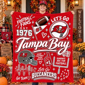 May include: A red fleece blanket featuring Tampa Bay Buccaneers football graphics, including a helmet, a football, a goalpost, the text 'Tampa Bay', 'Est 1976', 'Football Fans', 'Let's Go', 'Sundays Are For Football', and 'Go Team'.