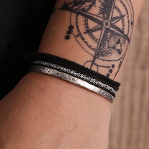May include: A stack of three bracelets. The bottom bracelet is silver-colored metal with a hammered texture. Above it is a black woven bracelet, and on top is a black rope bracelet. A compass rose tattoo is visible in the background.