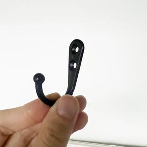Puede incluir: Un gancho de metal negro con un extremo redondeado y una placa de montaje con dos agujeros. El gancho se sostiene sobre un fondo blanco. Este peque&ntilde;o gancho curvado est&aacute; dise&ntilde;ado para colgar objetos.