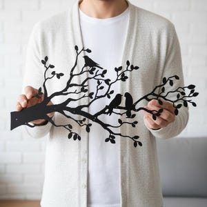 Puede incluir: Arte de pared de metal negro que representa una rama de &aacute;rbol con hojas y tres p&aacute;jaros. Los p&aacute;jaros est&aacute;n posados en las ramas. La obra de arte est&aacute; sostenida por una persona que lleva una camisa blanca y una rebeca gris claro.