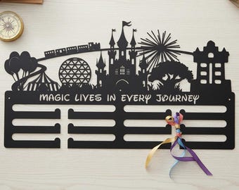 Puede incluir: Un colgador de medallas de metal negro para pared con la silueta de un castillo, un domo geodésico, una noria y un tren. El texto "MAGIC LIVES IN EVERY JOURNEY" está recortado sobre cuatro barras horizontales para colgar.