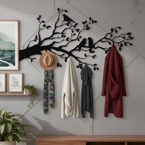 Puede incluir: Un perchero de pared de metal negro con forma de rama de &aacute;rbol con hojas y tres p&aacute;jaros posados. El perchero sostiene un sombrero fedora beige, una bufanda estampada, un su&eacute;ter blanco, una sudadera negra y un abrigo largo granate.