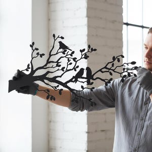 Puede incluir: Una pieza de arte mural de metal negro que presenta la silueta de una rama de &aacute;rbol con m&uacute;ltiples hojas y tres p&aacute;jaros posados en las ramas. El dise&ntilde;o es plano y est&aacute; cortado de una sola hoja de metal con acabado negro mate.